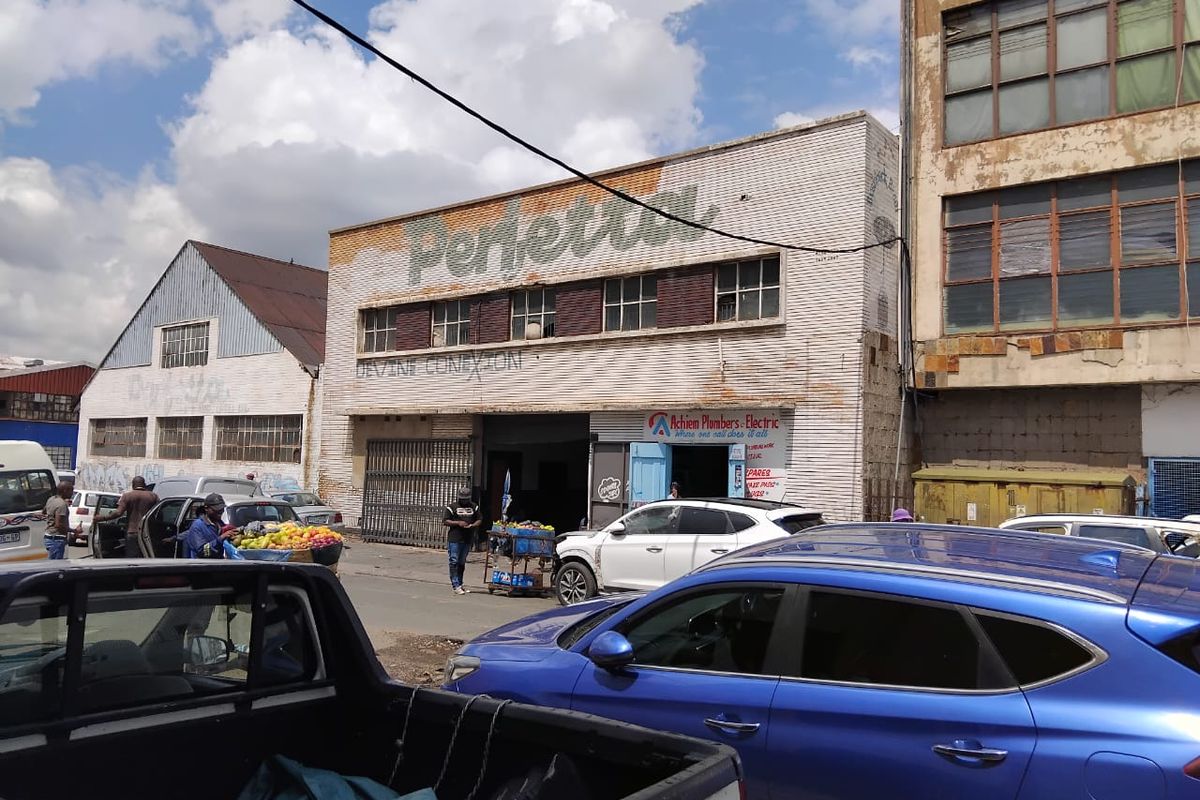 Street-level view of a commercial building featuring a brick facade, industrial windows, and metal security gates in a busy urban setting.