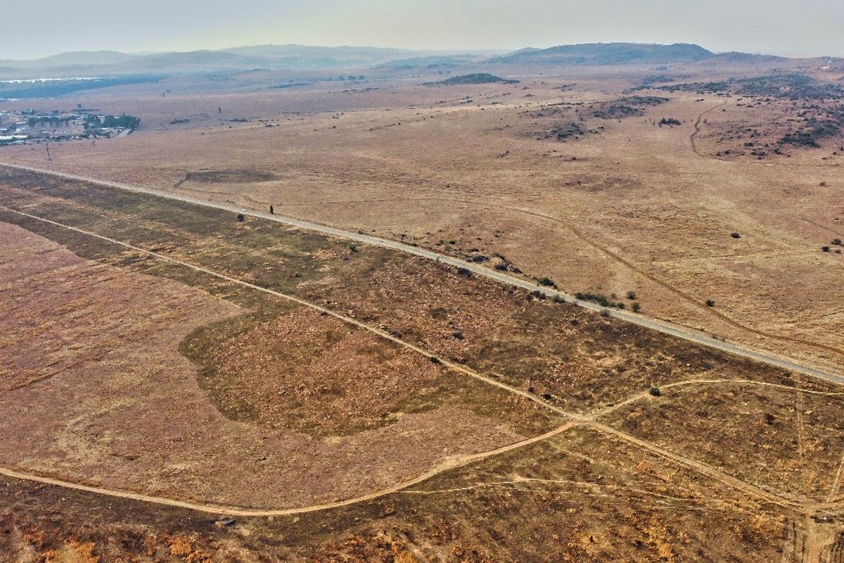 An aerial view of a large, undeveloped residential land parcel featuring natural terrain, dirt access tracks, and adjacent paved road frontage.