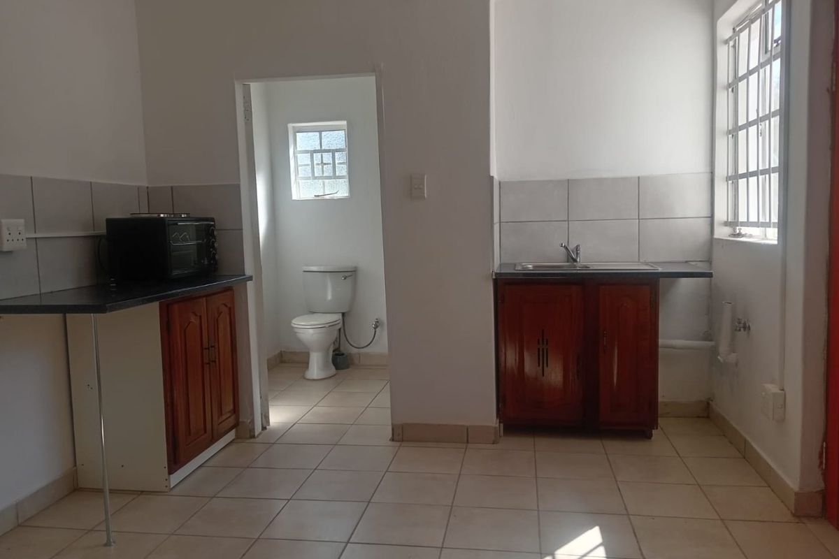 A functional kitchen area featuring built-in wooden cabinetry, laminate countertops, and a tiled backsplash, with an adjacent bathroom visible through the doorway.