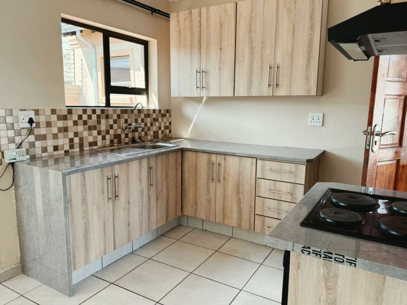 A functional kitchen featuring light wood-look cabinetry, stone-effect countertops, a mosaic tile backsplash, and built-in cooking appliances with ceramic floor tiling.