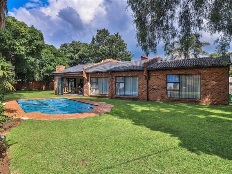 Rear view of a brick house featuring a swimming pool with a cover, a spacious lawn, and mature garden landscaping.