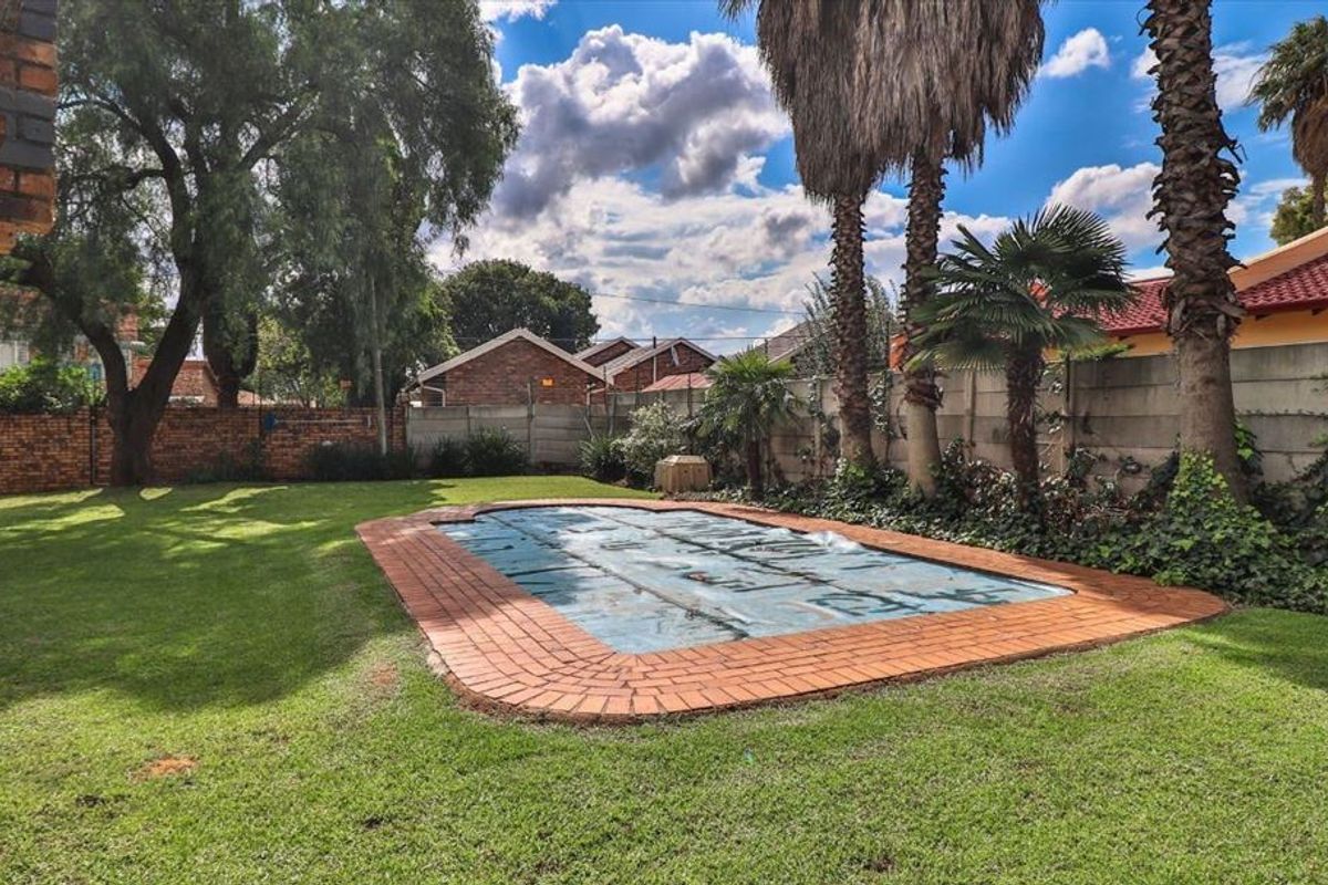 A spacious backyard featuring a swimming pool with a cover, surrounded by a well-maintained lawn, mature palm trees, and perimeter brick and concrete walls.