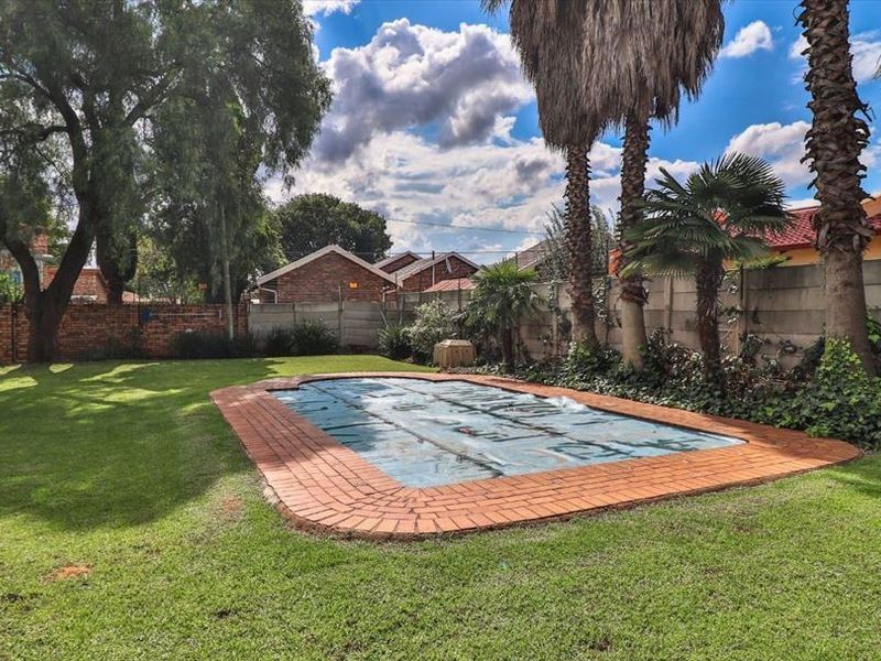 A spacious backyard featuring a swimming pool with a cover, surrounded by a well-maintained lawn, mature palm trees, and perimeter brick and concrete walls.