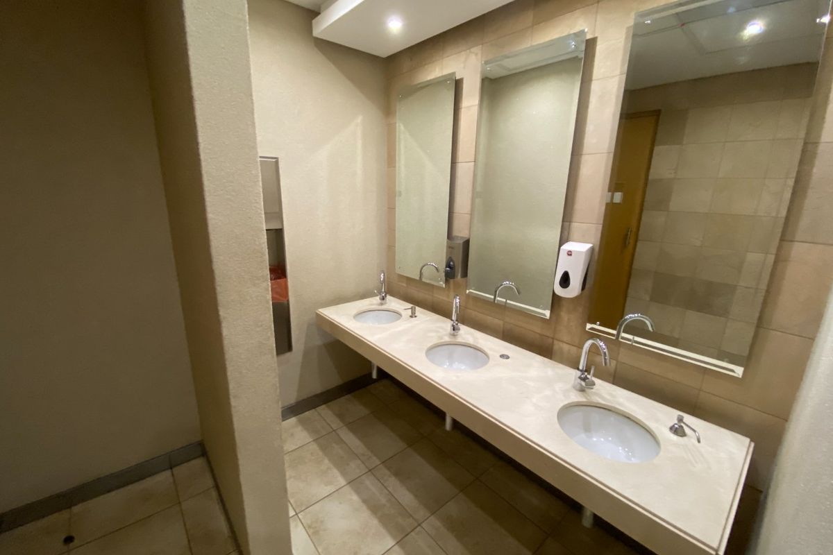 A clean commercial bathroom featuring a triple vanity with undermount sinks, chrome fixtures, and large wall-mounted mirrors with neutral tiled finishes.
