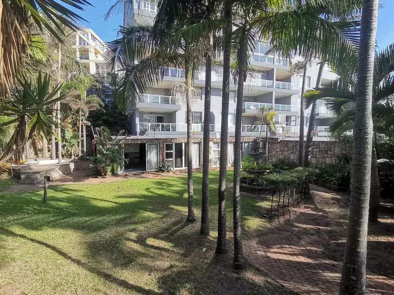 Rear view of the apartment complex featuring a landscaped garden, stone retaining walls, paved pathways, and private balconies with sliding door access.
