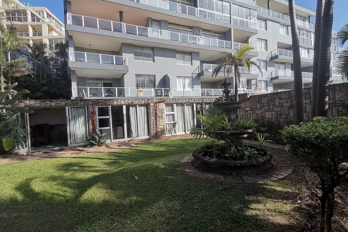 Rear view of a multi-story apartment complex featuring a landscaped garden, stone boundary wall, and a central water fountain.