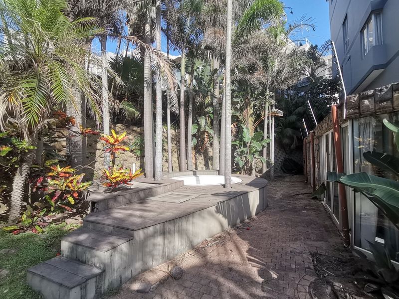 Outdoor area featuring a sunken spa set into timber decking, surrounded by mature palm trees and a paved walkway along the building exterior.