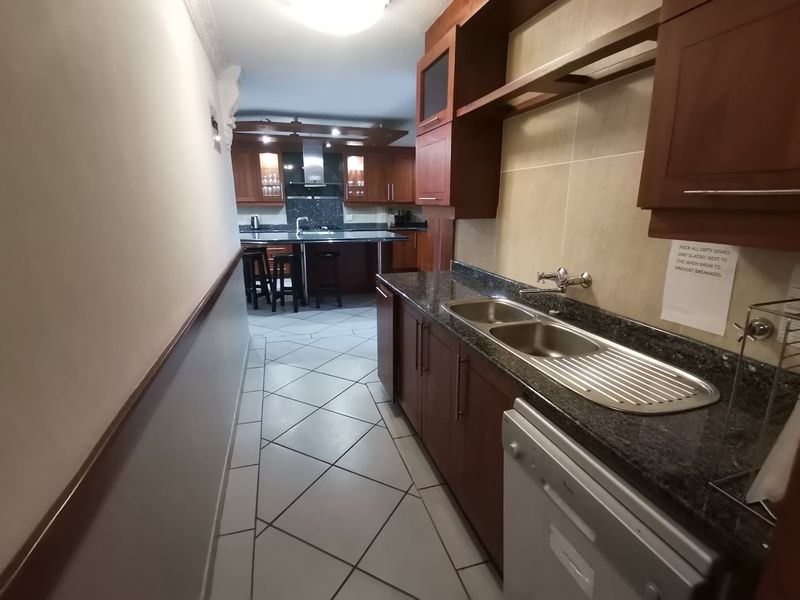A functional scullery area featuring granite countertops, wooden cabinetry, and a double stainless steel sink, leading into the main kitchen space.