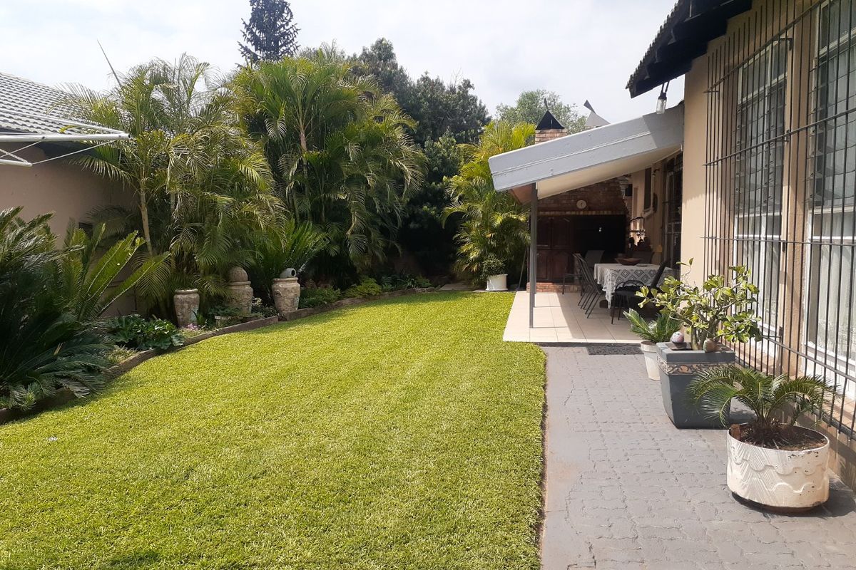 A well-maintained rear garden featuring a paved walkway, a covered patio area with a built-in braai, and established landscaping.