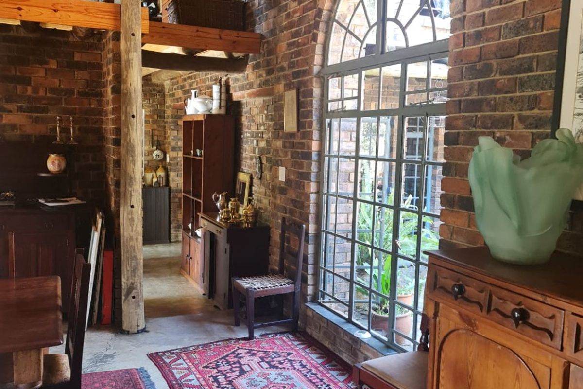 A rustic living area featuring exposed brick walls, a high thatched ceiling with timber beams, and a large arched window overlooking the garden.
