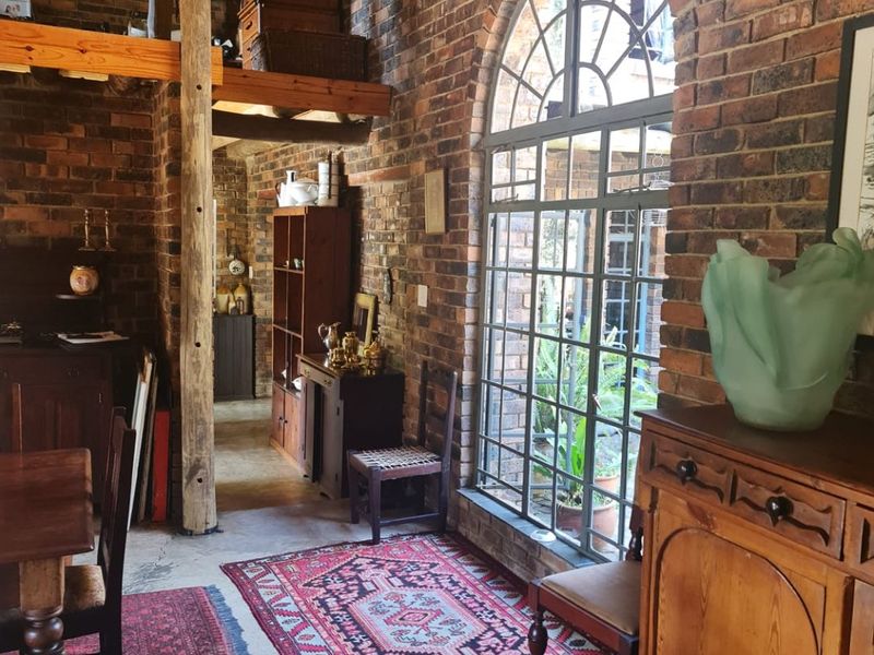 A rustic living area featuring exposed brick walls, a high thatched ceiling with timber beams, and a large arched window overlooking the garden.