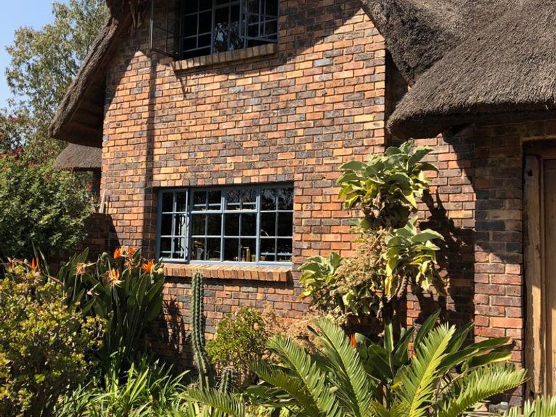 A charming farm-style residence featuring a traditional thatched roof, classic face brick exterior, and steel-framed windows surrounded by established garden landscaping.