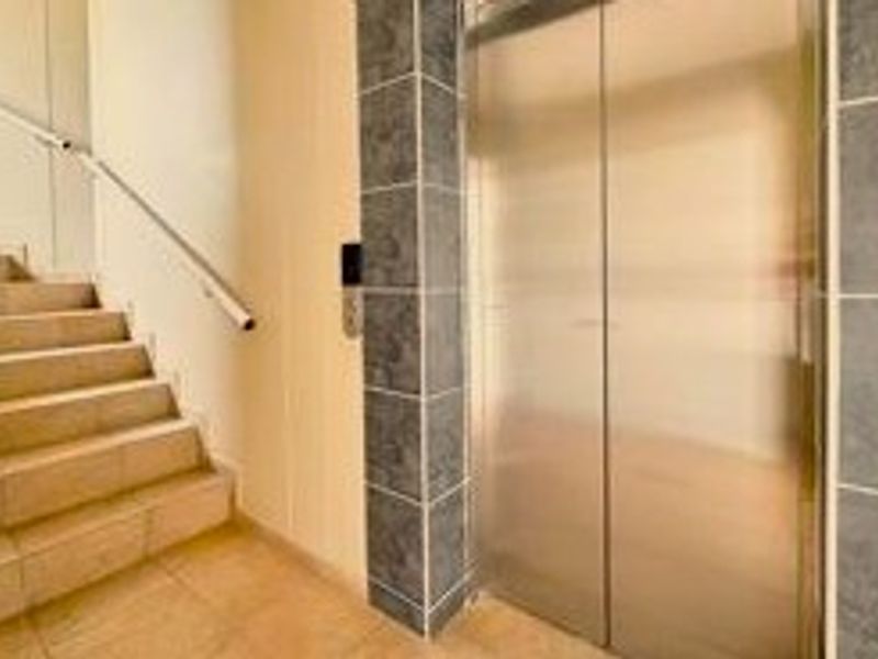 A clean apartment building hallway featuring a stainless steel elevator, tiled flooring, and a staircase with a metal handrail.