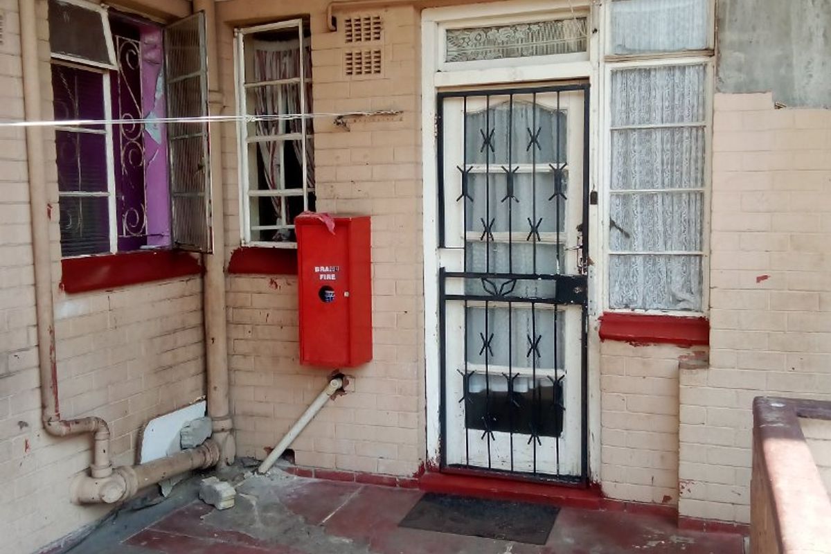 Exterior view of a commercial building entrance featuring a security gate, fire hose reel, and weathered brick facade with visible signs of wear.