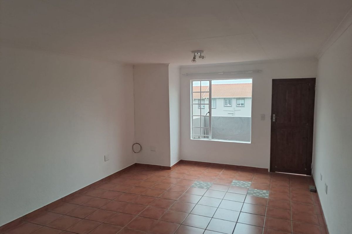 An empty living room featuring terracotta floor tiles, a large window, and a wooden door leading to the exterior.