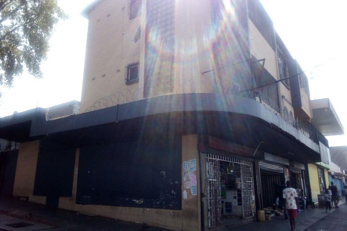 A commercial building exterior featuring a corner facade with roller shutter doors, security gates, and razor wire fencing along the upper levels.