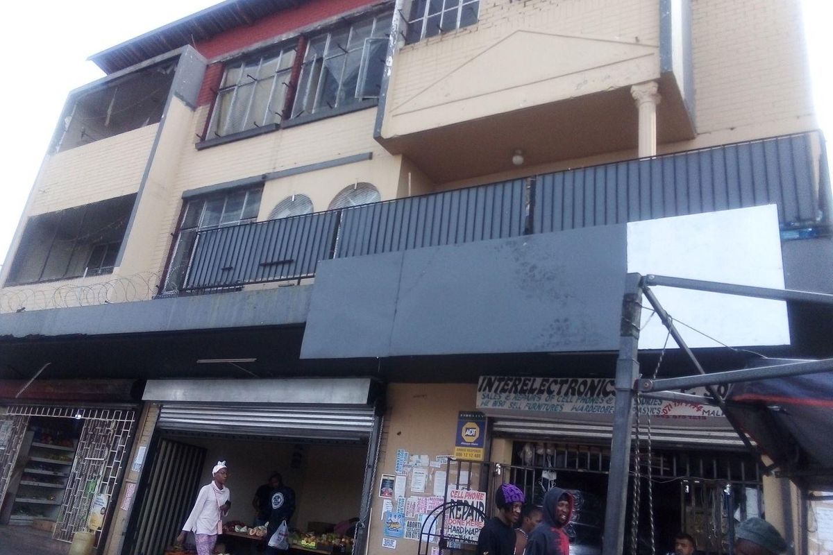 A multi-story commercial building facade featuring ground-level retail units with metal roller shutters and upper-level balconies with security railings.