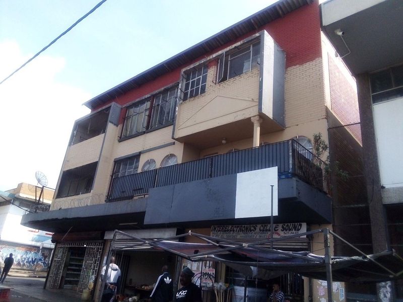 Street-level view of a multi-story commercial building featuring a brick facade, balcony with metal railing, and ground-floor retail storefronts.