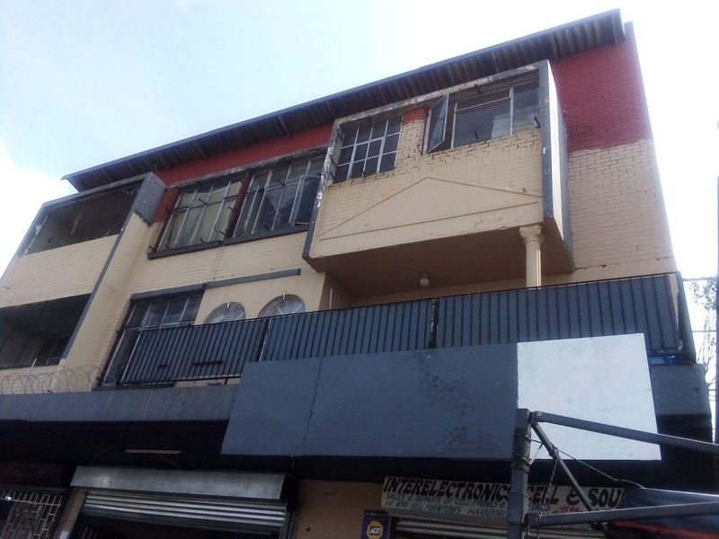 Front elevation of a multi-story commercial building featuring brick facade, metal balcony railings, and ground-level security roller shutters.