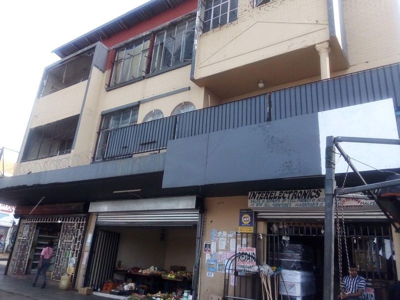 A multi-story commercial building facade featuring ground-level retail shopfronts with metal security shutters and upper-level balcony access.