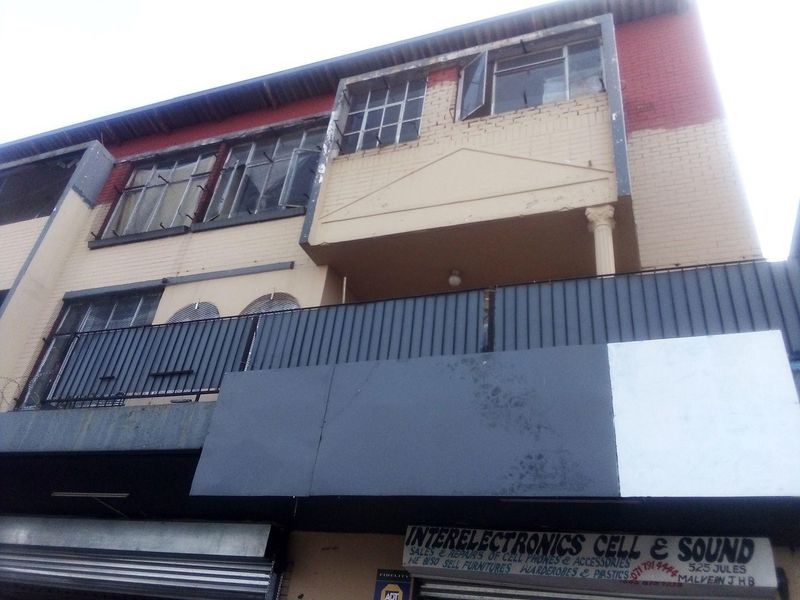 Front elevation of a commercial building featuring a ground-level storefront with security shutters and an upper-level balcony with steel railings.