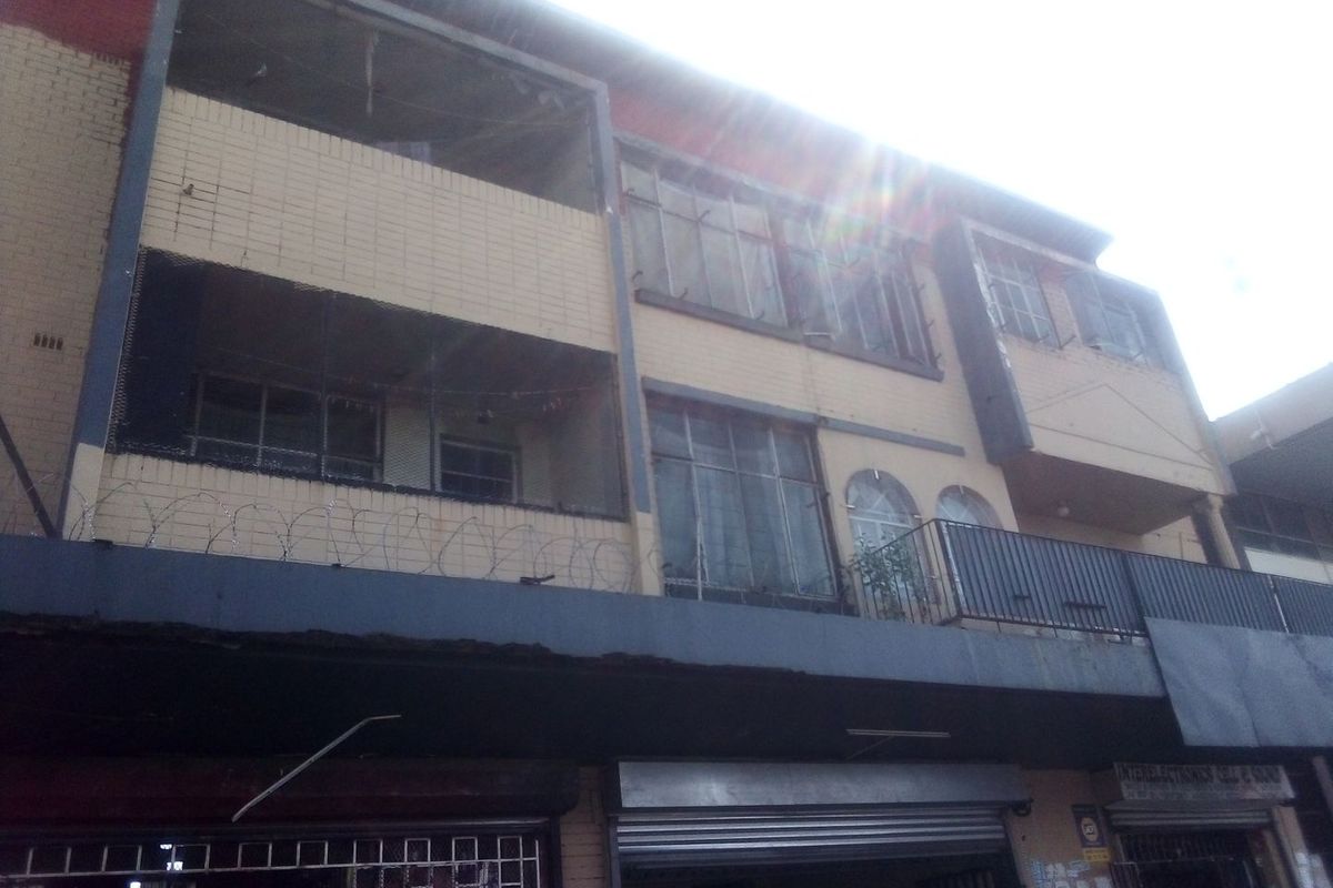 Exterior view of a multi-story commercial building featuring brick facade, balconies with metal railings, and ground-level security shutters.