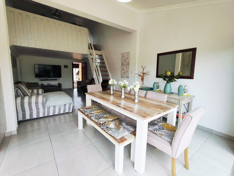 An open-plan living and dining area featuring a mezzanine level with a staircase, neutral tiled flooring, and a bright, airy layout.
