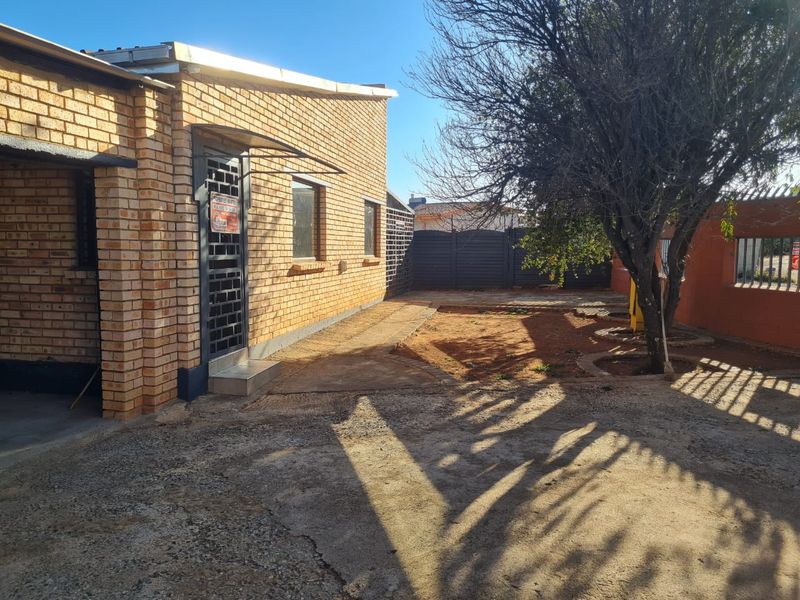 Rear exterior view of a face brick house featuring a paved walkway, security gate, and a spacious yard area with perimeter fencing.