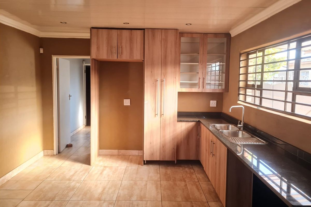 A clean, empty kitchen featuring wood-finish cabinetry, granite countertops, a double sink, and tiled flooring with recessed lighting.