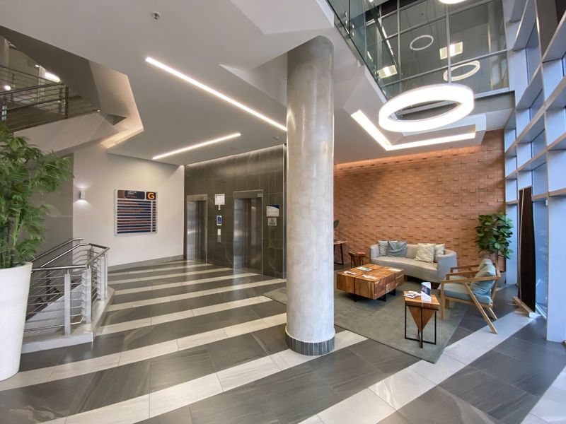 Modern commercial building lobby featuring a high-ceiling atrium, elevator access, exposed brick wall, and contemporary tiled flooring with integrated lighting.