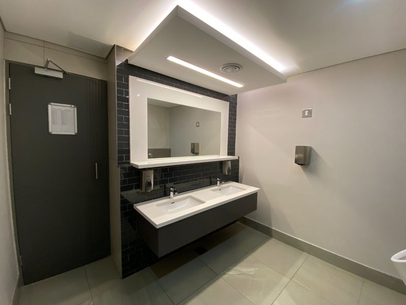 Modern commercial bathroom featuring a sleek double vanity with subway tile backsplash, integrated mirror, and recessed ceiling lighting.