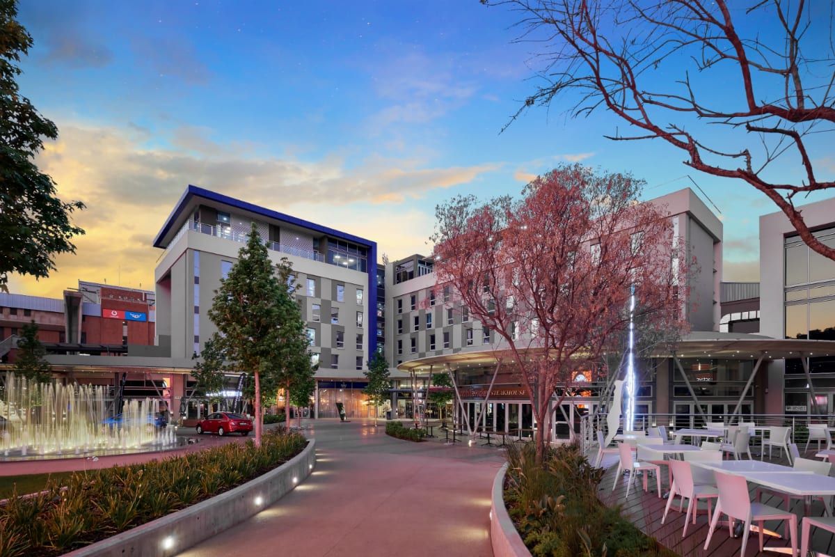 A well-maintained commercial plaza featuring a central fountain, landscaped gardens, paved walkways, and modern building facades at dusk.
