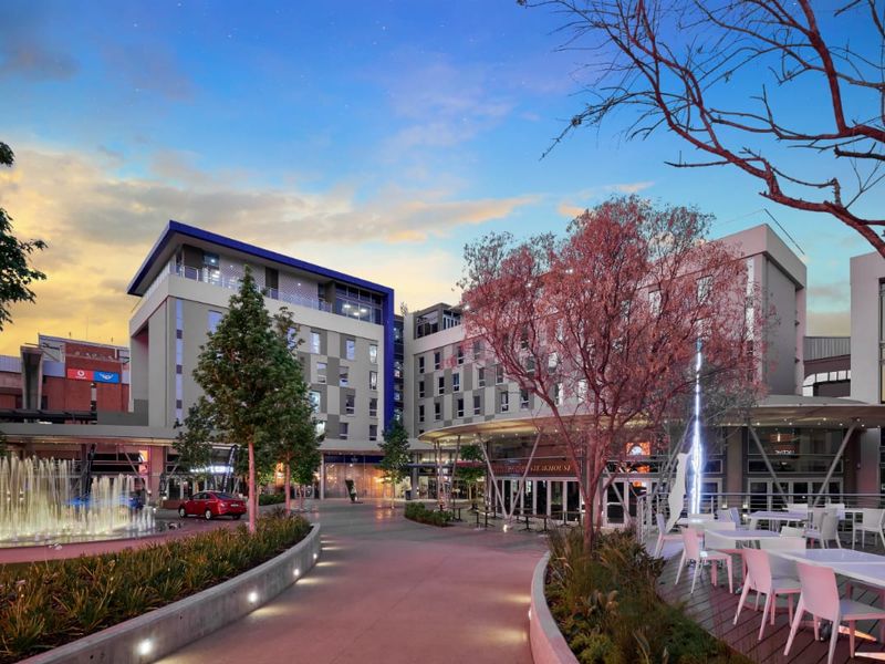 A well-maintained commercial plaza featuring a central fountain, landscaped gardens, paved walkways, and modern building facades at dusk.