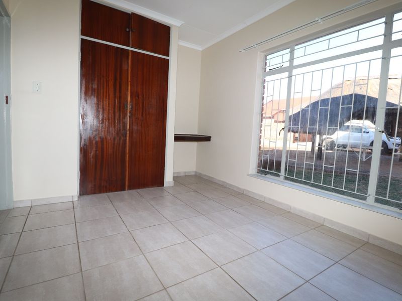 A bright, empty bedroom featuring neutral floor tiles, built-in wooden cupboards, and large windows fitted with security bars.