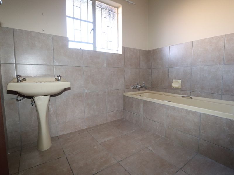 A dated bathroom featuring a pedestal basin, a built-in bathtub, and neutral wall and floor tiling with a security-barred window.