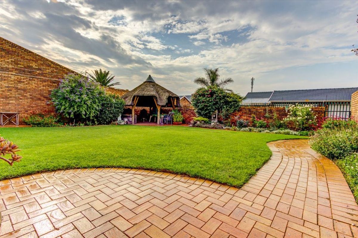 A well-maintained rear garden featuring a paved walkway, lush lawn, and a traditional thatched lapa enclosed by brick boundary walls.