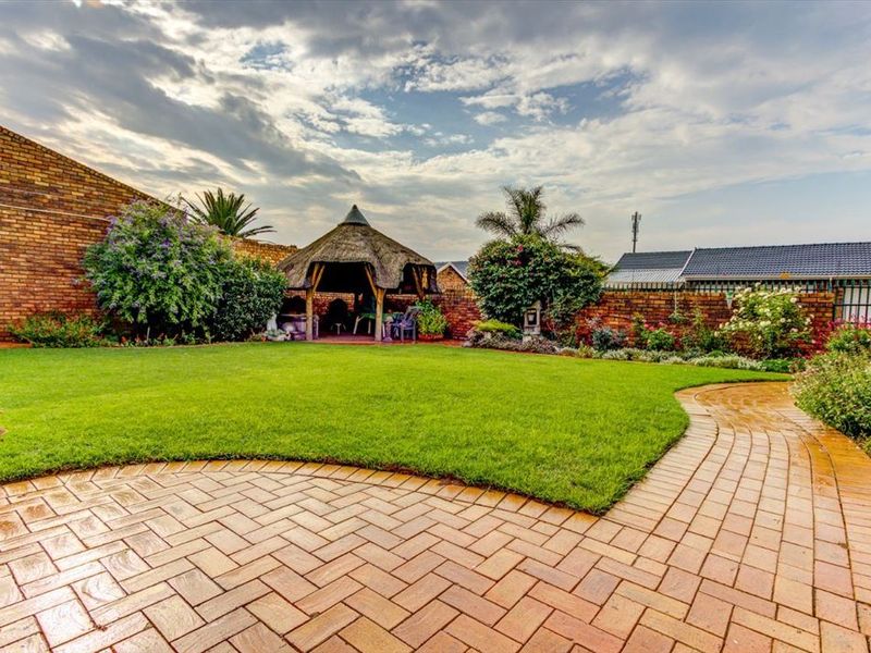 A well-maintained rear garden featuring a paved walkway, lush lawn, and a traditional thatched lapa enclosed by brick boundary walls.