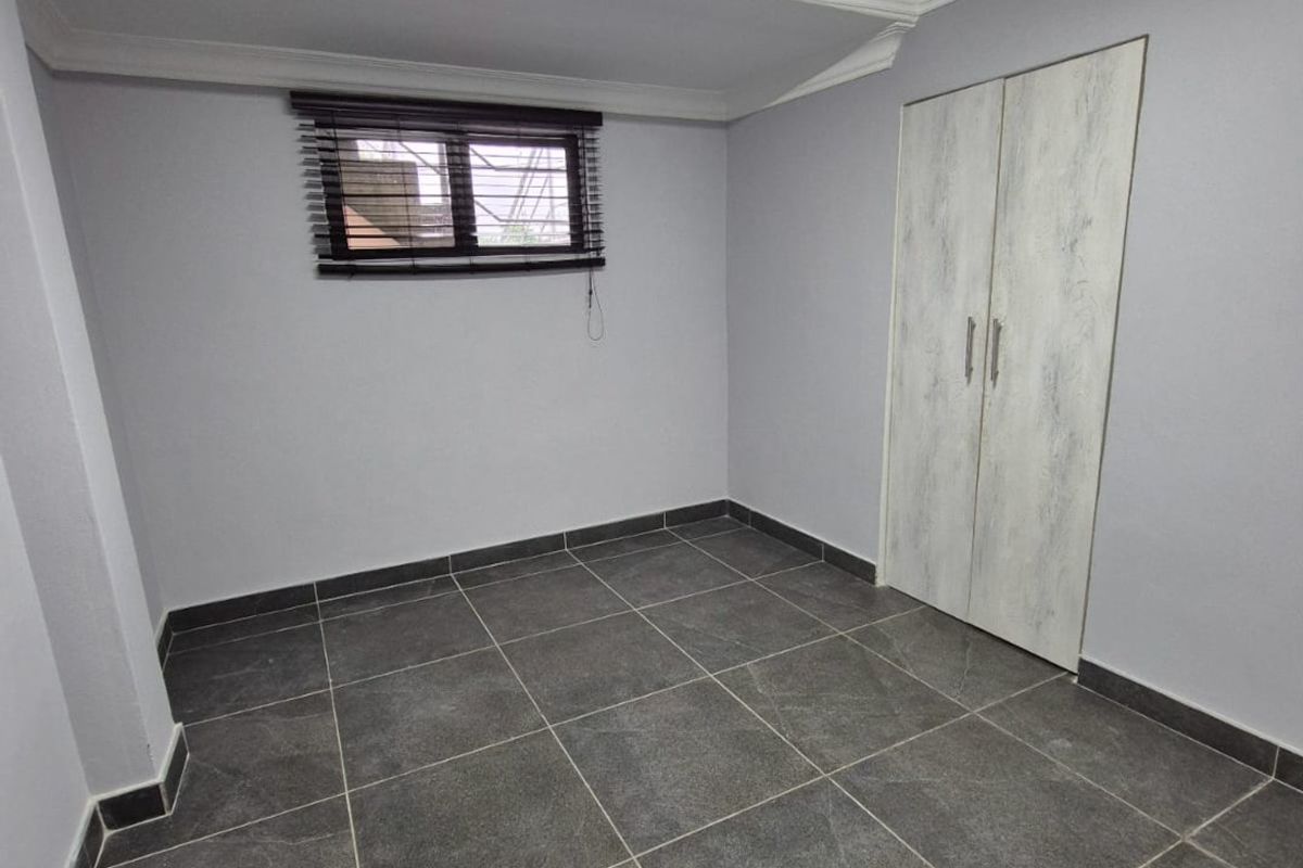 A clean, empty bedroom featuring modern dark grey floor tiles, neutral walls, a built-in wardrobe, and a window with security bars.