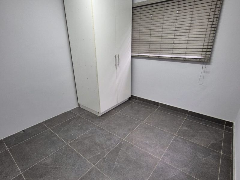 A clean, empty bedroom featuring modern dark grey floor tiles, a built-in white wardrobe, and neutral grey walls with fitted window blinds.