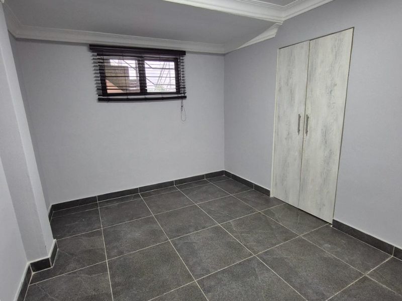 A clean, empty bedroom featuring modern dark grey floor tiles, neutral walls, a built-in wardrobe, and a window with security bars.