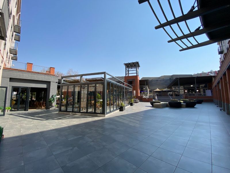 A spacious commercial courtyard featuring modern tiled flooring, a central glass-enclosed structure, and surrounding brick building facades.