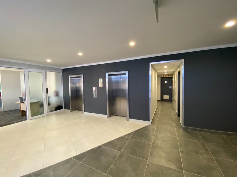 A modern commercial building lobby featuring stainless steel elevators, tiled flooring, and glass-partitioned office access.