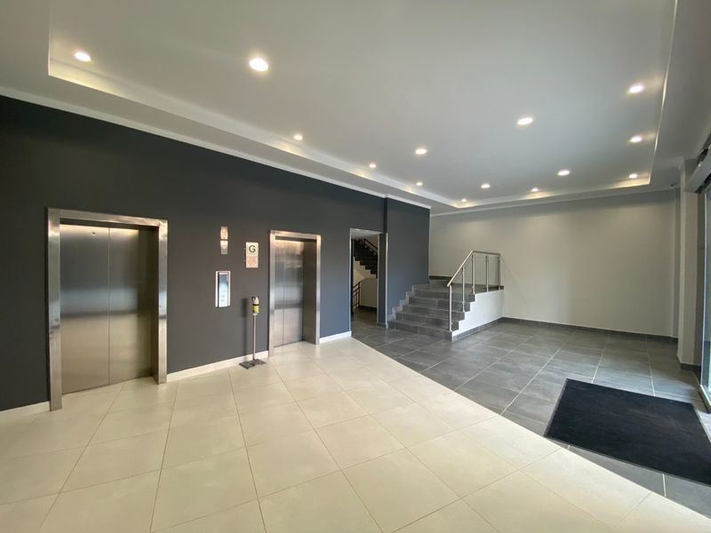Modern commercial building entrance lobby featuring dual elevators, tiled flooring, recessed ceiling lighting, and a staircase with a stainless steel handrail.