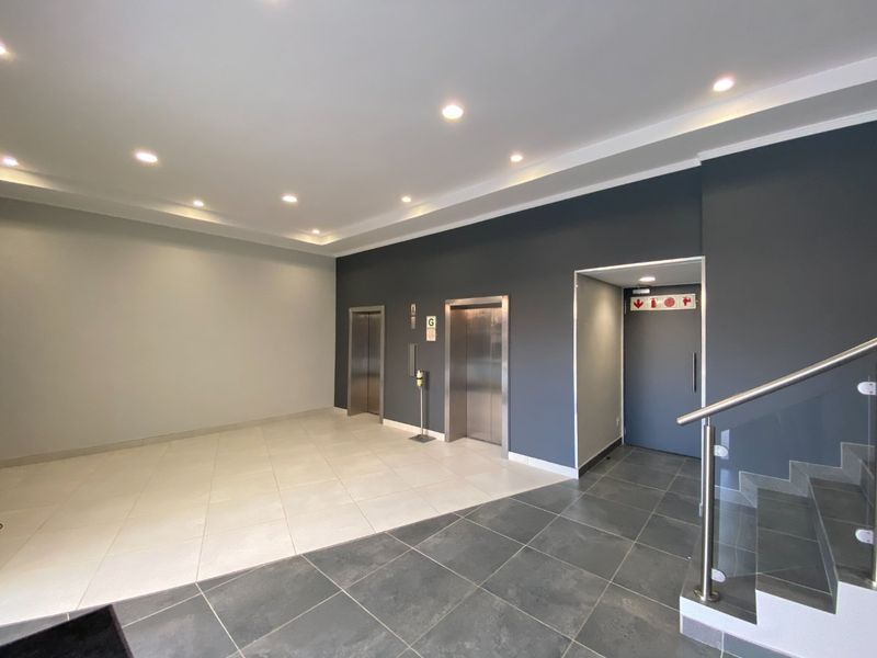 Modern commercial building lobby featuring stainless steel elevators, tiled flooring, recessed lighting, and a glass balustrade staircase.