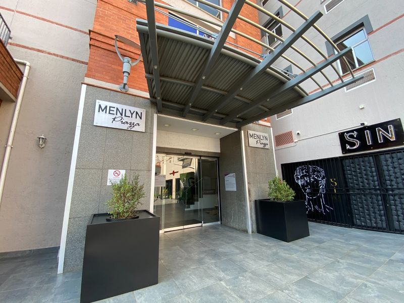 Main entrance to Menlyn Piazza featuring a modern metal awning, glass sliding doors, and stone-clad facade with professional signage.