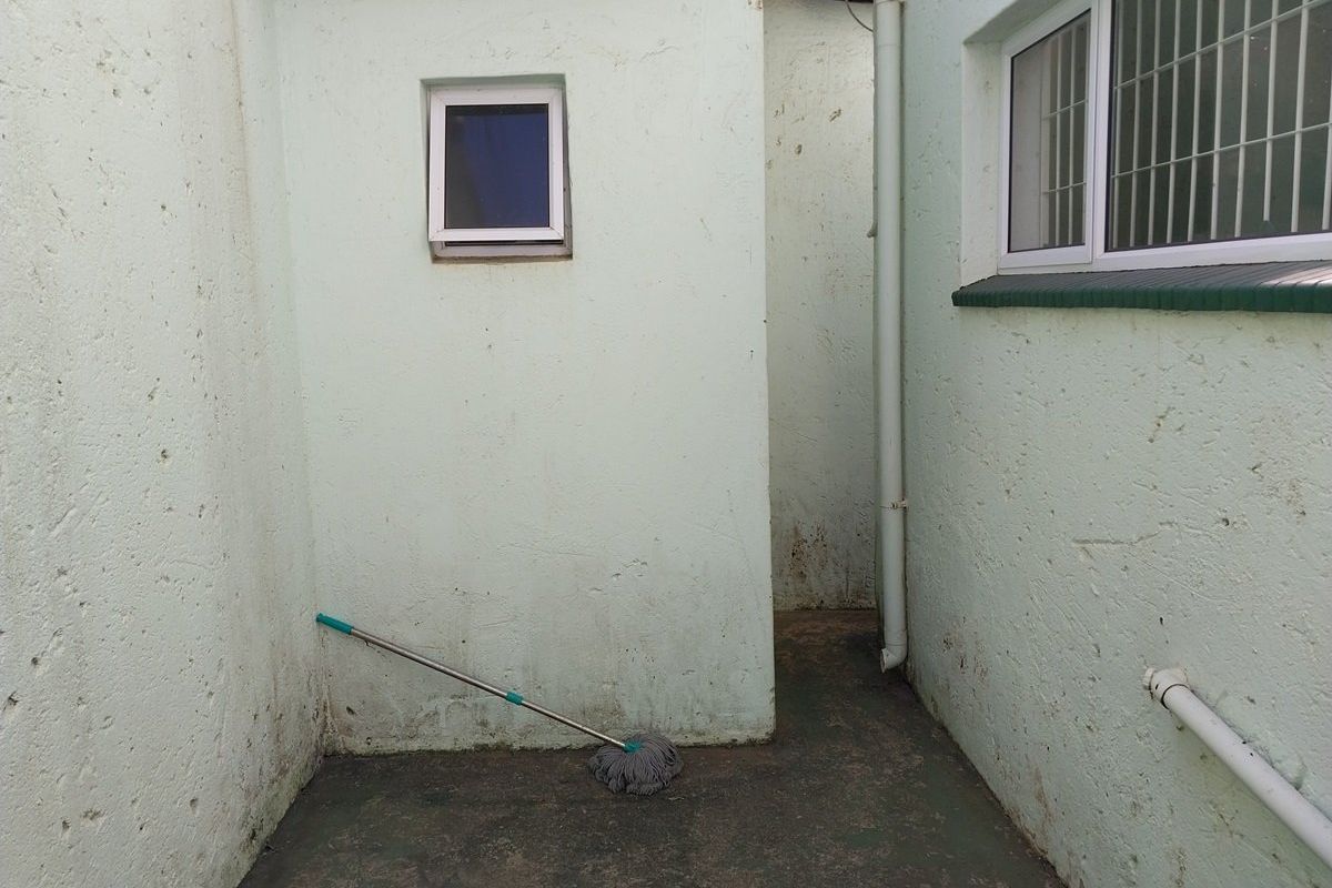 A narrow exterior rear service area featuring textured walls, security-barred windows, and a concrete floor, requiring maintenance and cosmetic updates.
