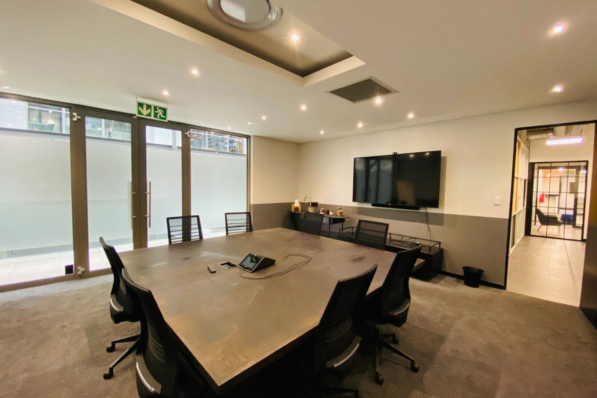 A professional commercial boardroom featuring a large central table, recessed ceiling lighting, carpeted flooring, and glass partition doors leading to an exterior area.