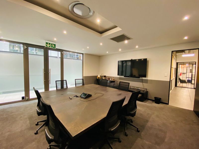 A professional commercial boardroom featuring a large central table, recessed ceiling lighting, carpeted flooring, and glass partition doors leading to an exterior area.