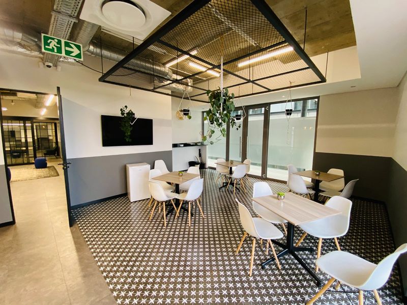Modern open-plan commercial breakout area featuring patterned floor tiles, exposed industrial ceiling with suspended mesh detail, and large glass doors providing natural light.