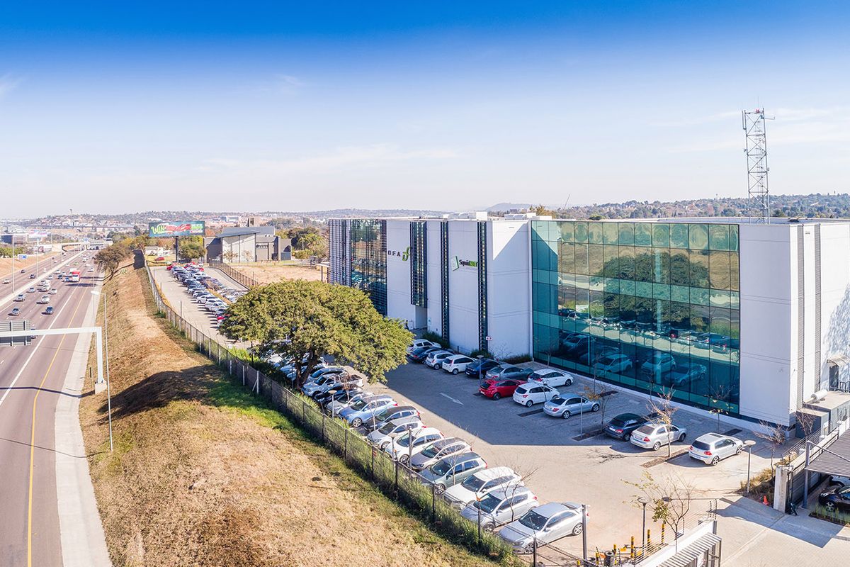 Aerial view of a modern commercial office building featuring a glass facade, extensive paved parking, and proximity to a major highway.